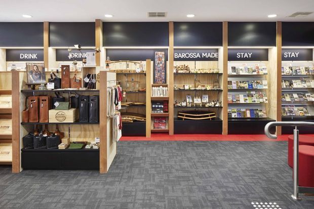 Interior view of a branded retail and visitor information space, with wall-mounted shelving and display fixtures stocked with books, merchandise, souvenirs, and local products. The fit-out uses timber-framed shelving, dark feature panels, integrated lighting, a red carpet strip, and grey patterned carpet flooring. A curved metal-and-glass balustrade or queue barrier is visible in the foreground.