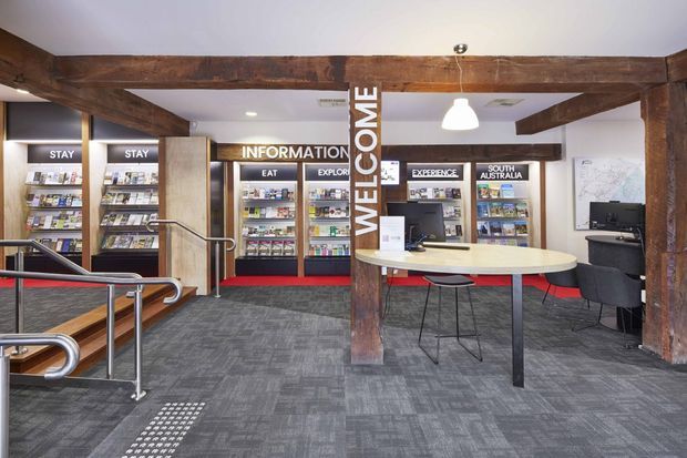 A bright, modern visitor information and reception area with exposed dark timber beams and posts, grey carpet flooring, built-in display shelving filled with brochures, a central welcome sign, a curved light-coloured reception counter with stools, lounge seating, a computer workstation, and accessible ramp railings. The space has a practical commercial fit-out with warm timber accents and clear wayfinding signage.