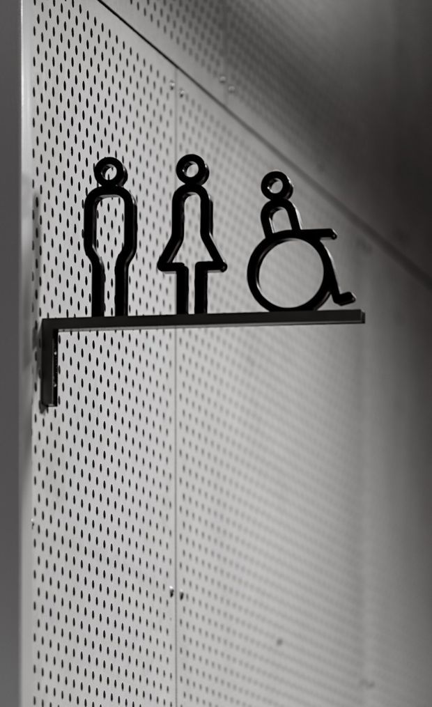 Close-up monochrome image of a minimalist accessibility sign showing male, female, and wheelchair symbols mounted on a narrow black shelf. The sign is set against a perforated metal wall panel, creating a clean, modern commercial interior detail.