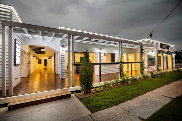 Exterior dusk view of a low-rise building with a covered veranda and open-air circulation area. The facade combines white horizontal weatherboard cladding, dark timber posts and rails, exposed white roof framing, warm timber decking, and illuminated glazed doors and windows. A landscaped lawn, planted garden bed, and concrete pedestrian path sit in front, while a long warmly lit hallway is visible through the open veranda.