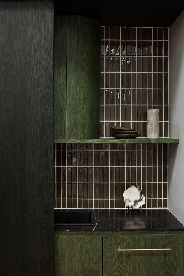 A moody, sophisticated built-in bar or beverage station with dark green wood-grain cabinetry, a black polished stone benchtop and integrated sink, and a glossy dark rectangular-tile backsplash. Open shelving displays bowls and sculptural decor, with slim brass hardware adding a refined accent. The compact joinery is set against dark walls and a lighter side wall.