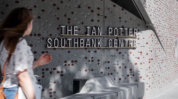 Photograph of the entrance or foyer wall at The Ian Potter Southbank Centre. A perforated grey concrete wall with recessed circular openings and a projecting white perforated panel frame illuminated lettering reading the centre's name. A person is walking past in the foreground, and a built-in concrete bench sits below the signage.
