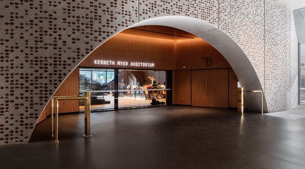 Interior entrance lobby to the Kenneth Myer Auditorium, featuring a dramatic arched opening, warm timber-lined soffit and wall panels, patterned perforated wall surfaces, dark flooring, brass-toned handrails, and glazed doors revealing the auditorium foyer beyond.