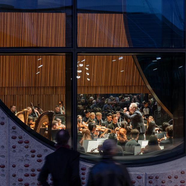 View through large dark-framed glazing into a concert hall, showing an orchestra performing beneath warm vertical timber acoustic wall panelling. Curved foreground architectural surfaces with circular red and dark insets and silhouetted visitors add depth to the scene.