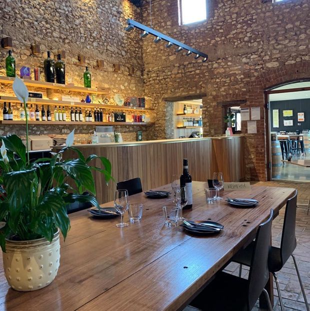 A rustic restaurant or wine-bar interior with a large timber dining table set with dark plates, wine glasses and a bottle. Behind it is a timber-fronted bar with open shelves displaying bottles, set against exposed irregular stone and brick walls. Warm integrated shelf lighting, a suspended linear track light and a high window create a relaxed, characterful atmosphere. An open doorway reveals additional dining or bar seating beyond.