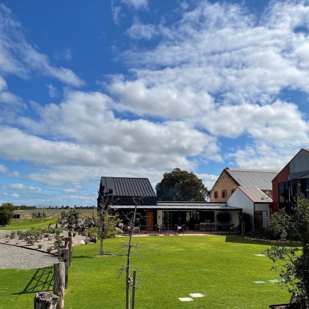 A rural homestead viewed from a landscaped lawn, featuring a contemporary dark-clad single-storey pavilion connected to traditional brick and metal-roofed buildings. The property includes an expansive manicured grass area, gravel paths, young orchard trees, garden beds, and agricultural fields beyond, under a bright partly cloudy sky.
