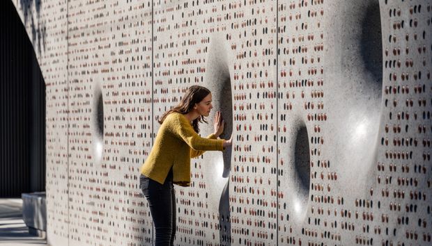 A person interacts with a sculptural perforated concrete wall composed of tall panels filled with small repeated openings and occasional larger recessed voids. Strong directional lighting creates dramatic shadows across the textured surface, with a dark vertical facade element and paved area visible at the edge.