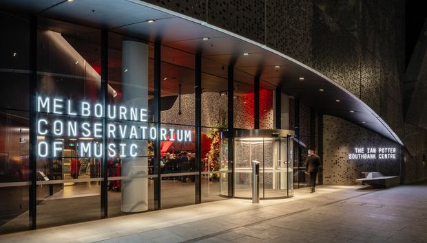 Night-time exterior view of the Melbourne Concert Hall at the Ian Potter Southbank Centre, featuring a curved perforated dark facade, extensive floor-to-ceiling glazed entry frontage, illuminated signage, a revolving entrance door, and a covered pedestrian forecourt.