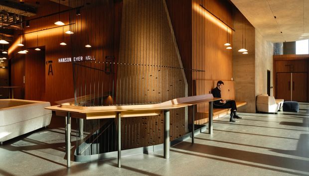 A warm, contemporary institutional lobby or entry hall with a large curved reception desk surrounding a stairwell, vertical timber wall panelling, exposed concrete surfaces, pendant lighting, and a glazed or open entrance area. The space is identified as Hanson Dyer Hall and includes a seated person near the circulation area.