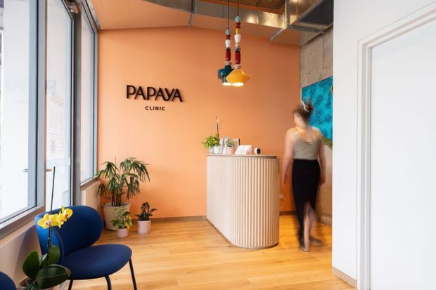 A compact, warmly styled clinic reception area with a curved fluted timber reception desk, peach-orange feature wall bearing the “PAPAYA CLINIC” sign, timber flooring, indoor plants, a blue upholstered lounge chair, colourful pendant lighting, large windows, and exposed ceiling services. A partially visible artwork and a person appear near the reception desk.