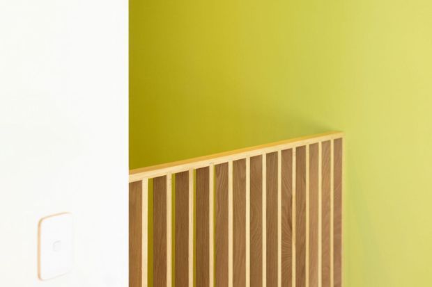 Close-up interior detail of a vertical timber slatted screen or balustrade against a vivid yellow-green painted wall, with a white wall and light switch visible at the left edge. The image focuses on fixed joinery and contrasting colour blocking.