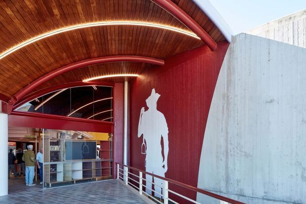 A covered entry or passageway to a contemporary commercial building, featuring a curved timber-lined ceiling with integrated linear lighting, red painted steel structure and wall panels, white metal handrails, a large white silhouette graphic, and a raw concrete wall. Glazed doors and a reception or interior space are visible at the far end.
