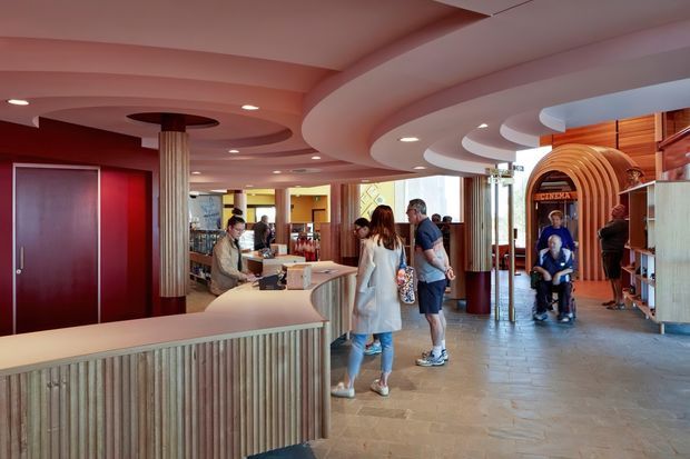 A lively contemporary hospitality reception or lobby with a sweeping curved front desk, ribbed vertical timber detailing, rounded layered ceiling soffits, recessed lighting, warm red and wood finishes, and stone-look flooring. Several visitors and staff occupy the open space, with a curved timber feature entrance or service portal visible at the rear.