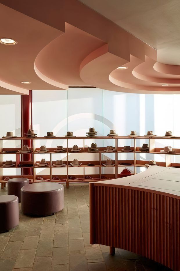 A stylish hat showroom with curved, sculptural terracotta ceiling forms, expansive translucent glazing, and warm timber display shelving filled with hats. A large rounded fluted timber counter dominates the foreground, complemented by cylindrical upholstered stools and a muted stone-tile floor. The space has a warm, polished, contemporary retail aesthetic.