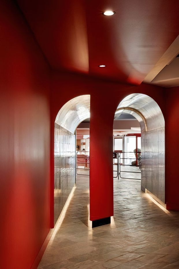 A dramatic interior corridor with deep red walls and ceiling, two rounded arched openings lined in reflective silver metal, and a pale irregular stone-tile floor. Recessed ceiling lights and concealed linear floor lighting emphasize the passage toward a brighter open interior beyond. The space has a bold, highly crafted contemporary character with strong geometric and metallic detailing.