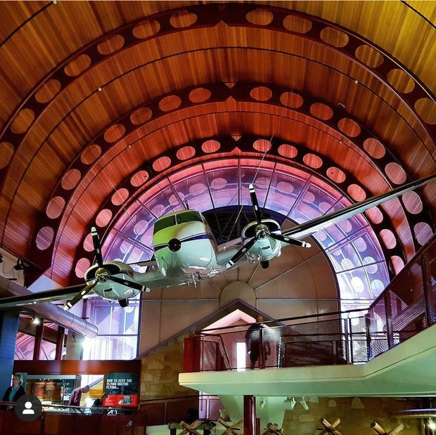 Interior of a dramatic aviation-themed commercial or museum space, featuring a suspended vintage twin-engine aircraft beneath a large curved timber barrel-vault ceiling. Layered arched timber ribs with oval cutouts, extensive glazing, mezzanine railings, and colourful accent lighting create a theatrical, industrial-exhibition atmosphere.