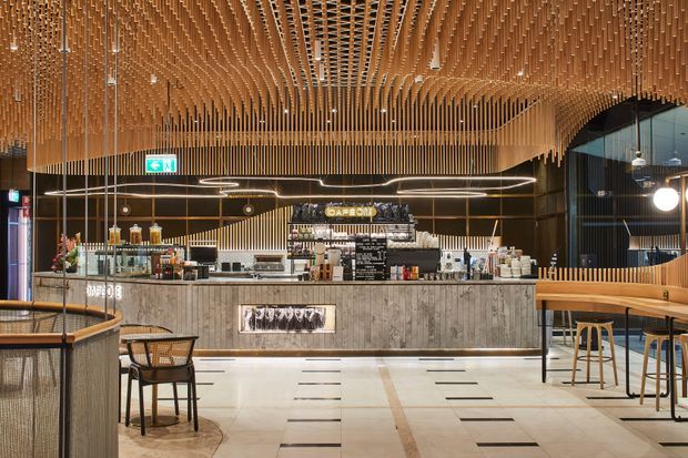 A sophisticated café or hospitality interior with a central service counter, display shelving, bar seating, and loose dining furniture. The space features a dramatic suspended timber-slat ceiling and matching vertical timber screening, dark walls and glazing, a textured grey concrete-look counter front, pale tiled flooring, and warm decorative lighting. The composition is refined and contemporary with strong linear detailing and an inviting atmosphere.
