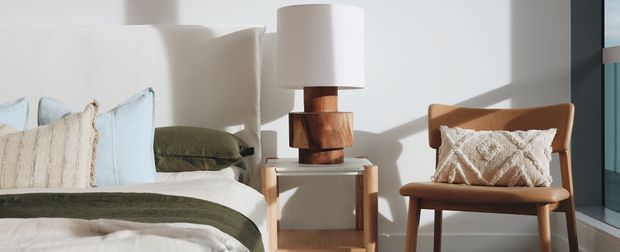 Bright, calm bedroom vignette featuring a neatly made bed with pale blue and cream pillows, an olive-green cushion and throw, a timber bedside table, a sculptural timber lamp with a white cylindrical shade, and a matching timber lounge chair with a textured cream cushion. Strong natural light enters from the right, creating crisp shadows on the white wall.