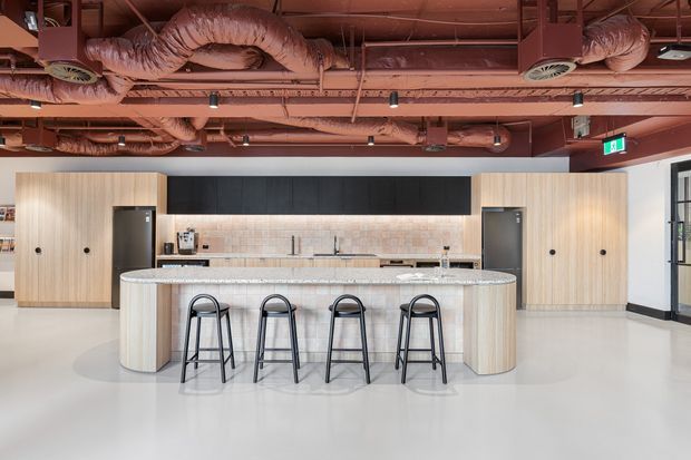 A spacious contemporary communal kitchen or hospitality-style break area with a large curved island and four black bar stools. The island has a pale stone benchtop and vertical light timber cladding, backed by light timber cabinetry, a pale tiled splashback, dark upper cabinetry, integrated appliances, and twin sinks. The exposed ceiling features prominent terracotta-painted ductwork, services, beams, and track lighting, creating an industrial character.