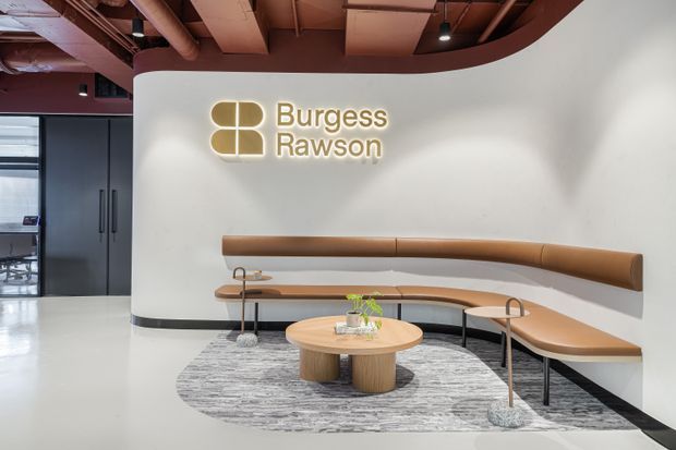 A contemporary commercial reception or waiting area featuring a curved white wall with illuminated Burgess Rawson signage, a built-in tan leather banquette wrapping around the corner, a round timber coffee table, small side tables, and a grey patterned rug. The space has a polished light floor, dark framed glazing and doors, exposed services, and a curved burgundy ceiling bulkhead.