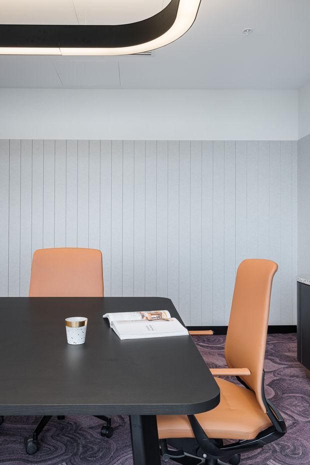 A contemporary meeting or conference room with a large rounded-edge black table, orange upholstered office chairs, pale blue-grey vertically panelled walls, patterned purple carpet, and a distinctive curved recessed ceiling light.