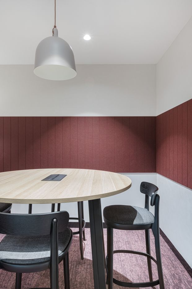 Contemporary dining area with a round pale timber table, dark upholstered bar-height chairs, a large white pendant light, and recessed ceiling lighting. The room features burgundy vertical wall panelling above a light grey lower wall treatment, with a muted patterned carpet underfoot.