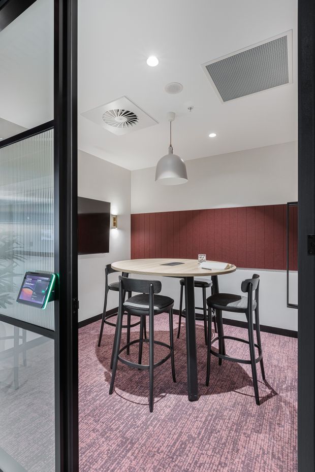 A compact contemporary meeting or breakout room viewed through a black-framed glazed doorway. The room contains a round light-wood table with four black bar-height chairs, a wall-mounted display, a red vertical-panel acoustic feature wall, and mauve patterned carpet. A grey pendant light and recessed ceiling services provide illumination, while textured glass and an access control panel are visible at the entrance.