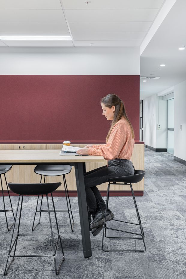 A bright contemporary commercial office breakout area with a long light-wood communal table, black bar stools and chairs, and a built-in timber cabinet beneath a large burgundy acoustic wall panel. The space has a white suspended ceiling with linear fluorescent lighting, grey patterned carpet tiles, and an open corridor or circulation area to the right.