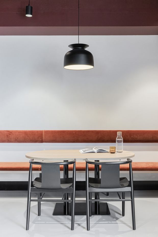 A minimalist dining nook with a light timber oval table, two dark-framed chairs, and a long rust-colored upholstered banquette against a pale wall. Black pendant lights hang from a dark reddish ceiling, while an open book, glass, and small tumbler sit on the table. The space has a refined, contemporary commercial-interior feel with strong geometric lines and restrained materials.