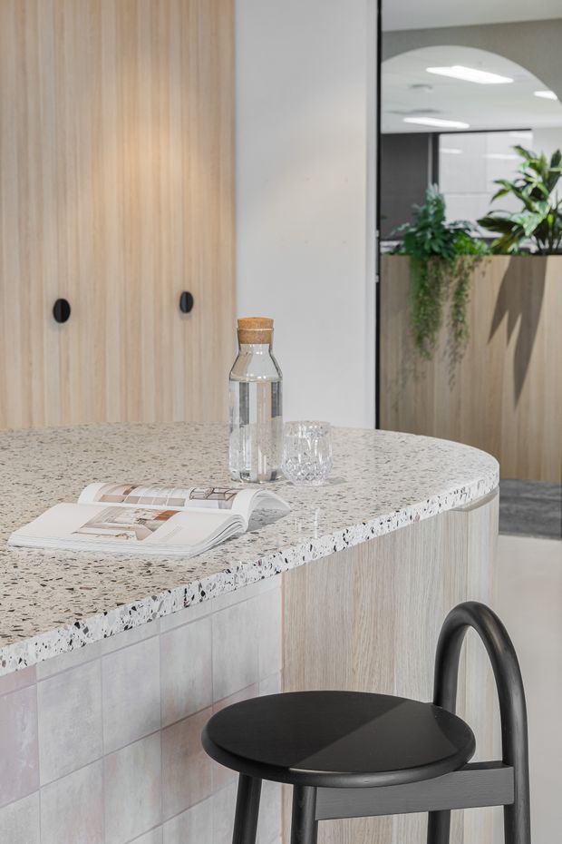 A bright, refined kitchen or hospitality-style counter area featuring a curved island with a terrazzo benchtop, pale timber cabinetry, and a tiled curved base. A black bar stool sits in the foreground, while a glass partition reveals additional pale timber joinery and indoor greenery beyond.