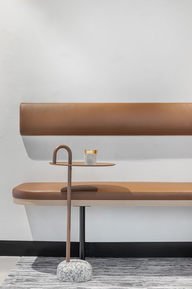 A minimalist interior vignette featuring a sculptural tan-brown leather upholstered bench with a matching horizontal backrest against a white wall. A slim bronze-toned pedestal side table with a circular tray top stands in front, holding a small cup. The bench has dark supports, and the scene sits on a muted grey patterned rug above a black skirting board.