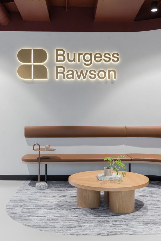 A refined commercial reception or waiting area featuring an illuminated Burgess Rawson logo on a light grey wall. A long built-in brown leather banquette runs along the wall, paired with a curved wood coffee table, a small side table and lamp, and a potted plant. The space has grey patterned carpet, dark skirting, and a warm timber ceiling with exposed services and pendant lighting.