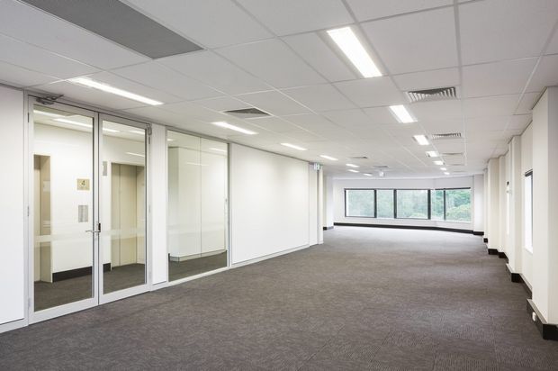A bright, vacant commercial office interior with an open-plan carpeted workspace, glazed office or meeting-room partitions along the left side, white walls, a suspended acoustic ceiling with recessed fluorescent lighting and HVAC vents, and a wide bank of windows at the far end. The space has a clean, functional corporate fit-out.