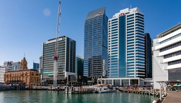 Waterfront urban cityscape featuring several modern high-rise commercial buildings, a construction crane, a historic stone building with a clock tower, and a marina or ferry wharf in the foreground under a clear blue sky.