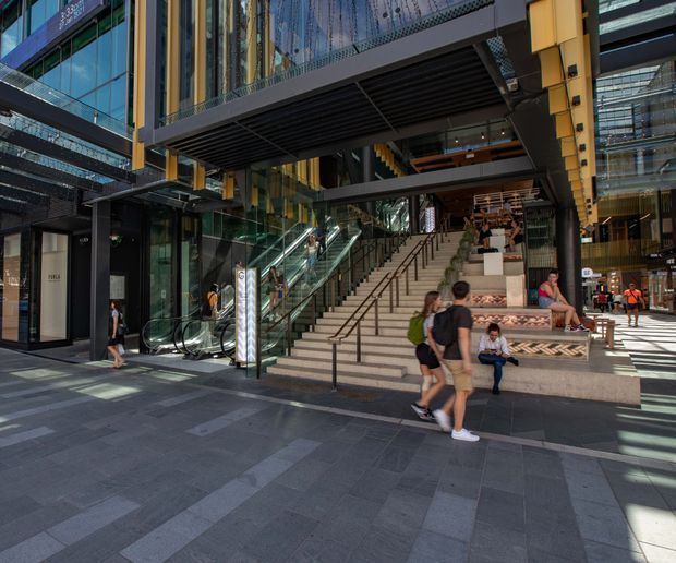 A large contemporary commercial atrium or urban retail precinct with broad stone steps, escalators, glass façades, dark metal canopies, and yellow structural framing. People are using the stepped seating and circulation areas, while shops and internal walkways are visible around the open multi-level space.