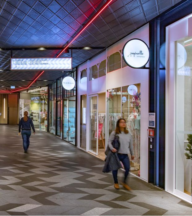 Interior view of a contemporary retail shopping arcade or commercial corridor, with multiple glazed shopfronts, illuminated signage, patterned geometric floor tiling, a decorative pressed-panel ceiling, and linear red accent lighting. The corridor has a polished, atmospheric fit-out with visible storefronts and pedestrians.