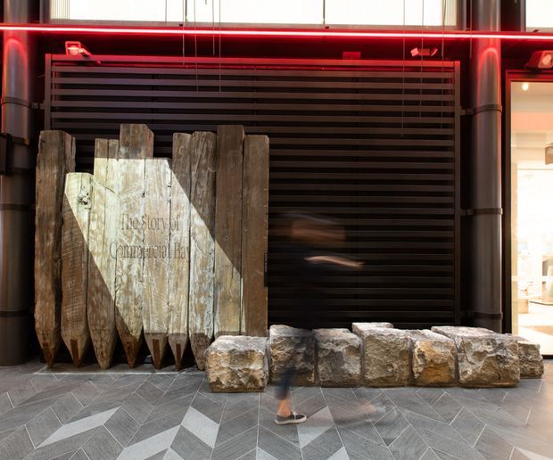 A dramatic commercial interior entrance or feature display with dark horizontal slatted wall panels, cylindrical dark columns, red linear accent lighting, and a sculptural installation made from weathered timber planks and rough stone blocks. The floor features a geometric grey patterned finish, while a bright adjoining retail or display area is visible at the right.