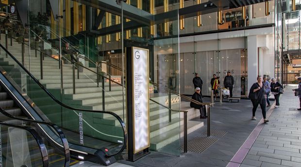 A contemporary multi-level commercial interior or transit-style public space with escalators, broad stairs, extensive glass balustrades, dark structural framing, and warm vertical timber or brass-toned accents. People occupy the circulation area, with retail or office frontage visible beyond the glazed partitions.
