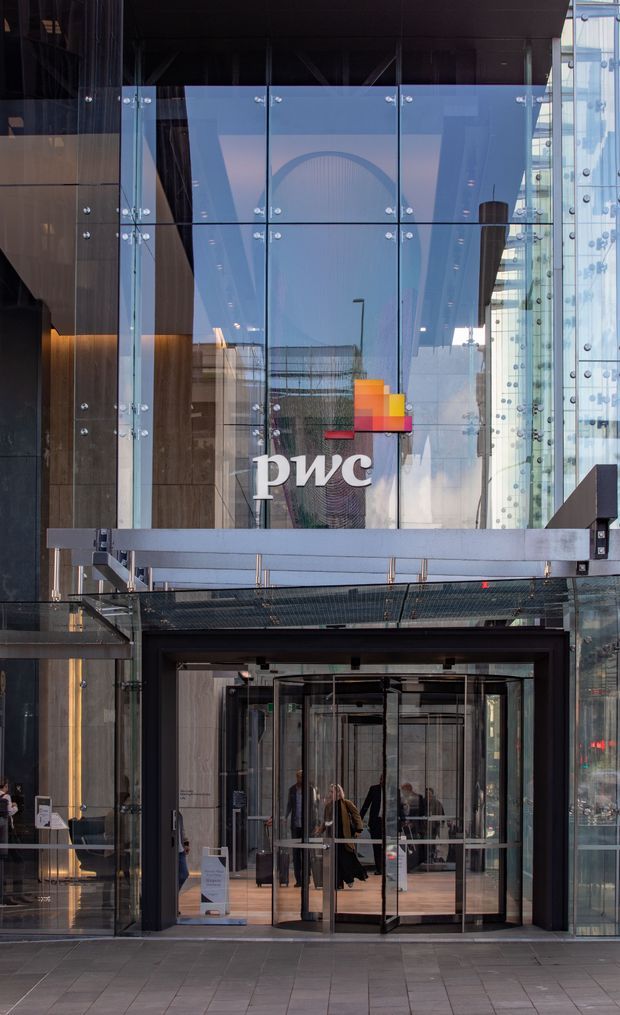 Exterior view of a contemporary commercial office building entrance, featuring a tall glazed curtain wall, exposed glass fixing points, metal framing, a projecting canopy, and revolving glass doors. The PwC logo is displayed prominently above the entrance.