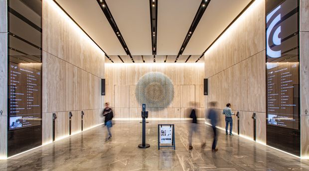 A polished contemporary commercial lobby or gallery entrance with floor-to-ceiling pale stone or wood-look wall panels, dark glossy display panels, linear recessed ceiling lighting, illuminated perimeter details, and a large circular feature artwork. Visitors are gathered around a freestanding information display, creating a refined institutional or corporate reception atmosphere.