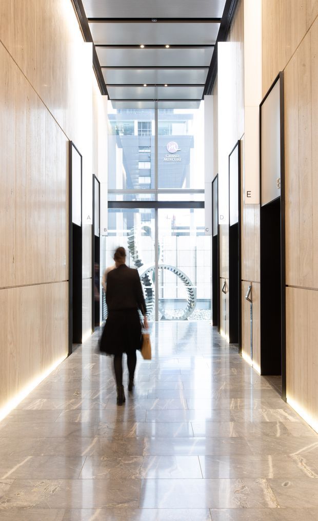 A bright, sophisticated commercial lobby or hotel corridor with pale timber wall panelling, polished stone flooring, black-framed illuminated directory panels, and a glazed roof with exposed dark framing. The corridor leads toward a full-height glazed entrance or atrium, with a person walking through the space and an urban building visible beyond.