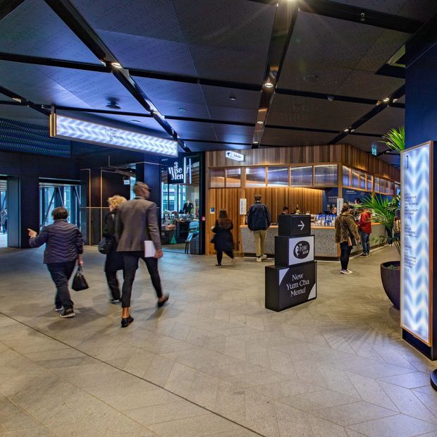 A contemporary commercial interior resembling a shopping centre or hospitality precinct, with a broad tiled concourse, illuminated retail and food outlets, timber-clad service counters, dark ceiling panels, dramatic blue accent lighting, freestanding signage, and several people moving through the space.