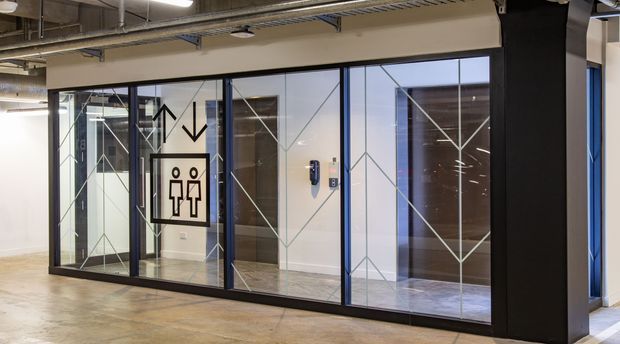 A modern commercial interior corridor with a glazed lift lobby and elevator doors. The space features black-framed full-height glass partitions, geometric line decals, polished concrete flooring, exposed services and ductwork, and an accessible restroom direction sign. The fit-out has an industrial, utilitarian character.
