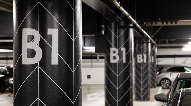 Interior view of a modern underground parking garage with black structural columns marked “B1” and decorated with white geometric line graphics. Parked cars, exposed ceiling services, bright strip lighting, and painted concrete surfaces are visible.