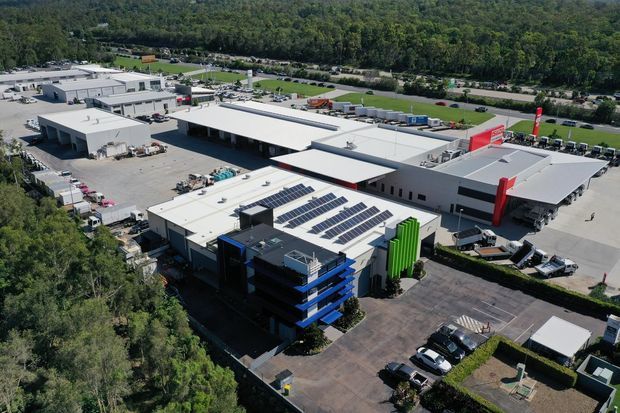 Aerial view of a large contemporary commercial and industrial complex with multiple low-rise buildings, expansive flat roofs, loading areas, parking, rooftop solar panels, and bold red and green facade elements. The site is surrounded by dense woodland and road infrastructure.