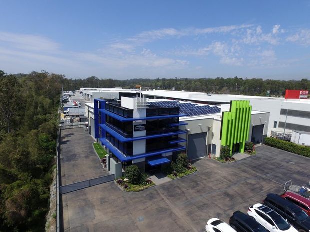 Aerial view of a contemporary commercial or industrial building complex with a striking blue-and-white multi-level office wing, rooftop solar panels, and a bright lime-green vertical facade feature. Large paved parking and service areas surround the building, with dense trees beyond the site.