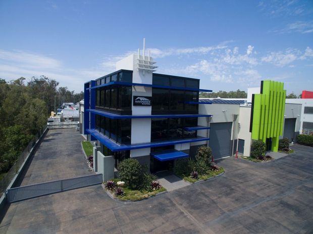 Aerial view of a contemporary multi-storey commercial building in an industrial or business park. The building features dark glazed curtain walls, bold blue horizontal edge bands, a white central rooftop structure, and a vivid lime-green vertical facade element, with paved access areas and landscaping around it.