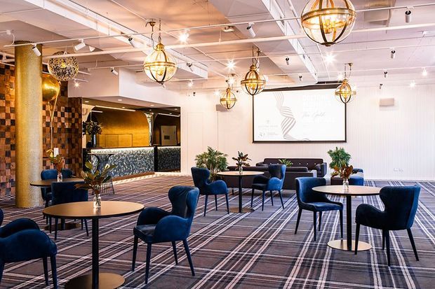 A stylish hospitality lounge or reception area with numerous round café tables and deep blue upholstered chairs arranged across a patterned carpet. The space features a reception or bar counter with decorative patterned cladding, timber and tiled wall accents, a large projection screen, exposed ceiling services, and multiple spherical brass-and-glass pendant lights.