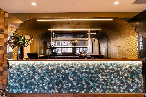 A stylish commercial bar or reception counter with a dramatic scalloped blue-green tiled front, dark timber side walls, and a large rounded metallic mosaic arch framing open shelving and back-bar equipment. Warm integrated linear lighting and reflective surfaces create a luxurious, atmospheric hospitality interior.