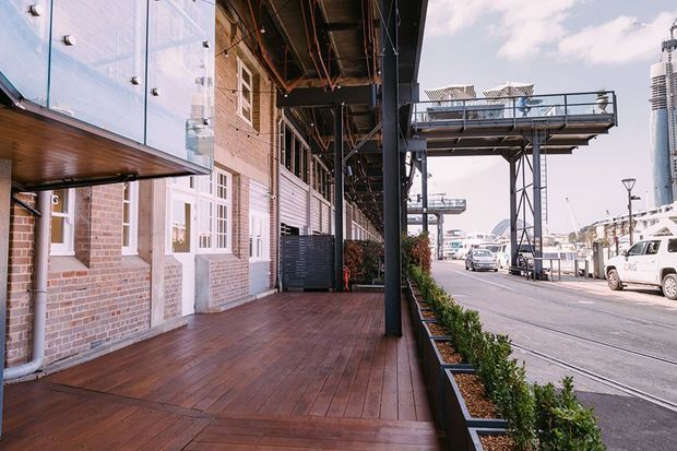 Exterior view of a brick commercial or mixed-use building along a wide urban street. The image shows a covered timber deck and pedestrian walkway, white-framed doors and windows, exposed dark steel columns and structure, a projecting glazed canopy, landscaped planter boxes, and elevated industrial-style structures across the street.